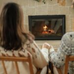Stylish couple in cozy sweaters relaxing and looking at fireplace with festive mantle on background of stylish decorated christmas tree with lights. Happy young family enjoying winter holidays