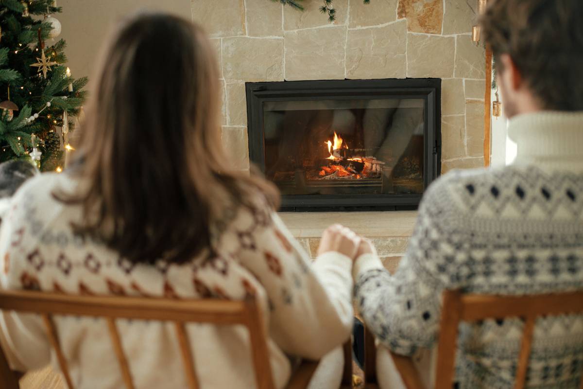 Stylish couple in cozy sweaters relaxing and looking at fireplace with festive mantle on background of stylish decorated christmas tree with lights. Happy young family enjoying winter holidays